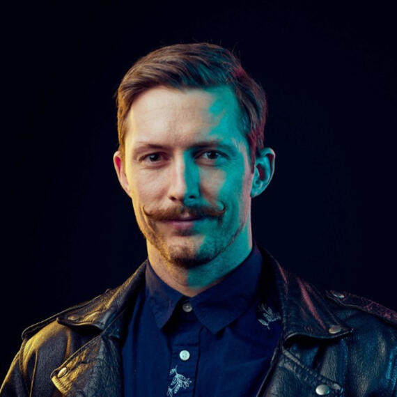 Picture of Robert Roche wearing a blue shirt, leather jacket, and a mustache.